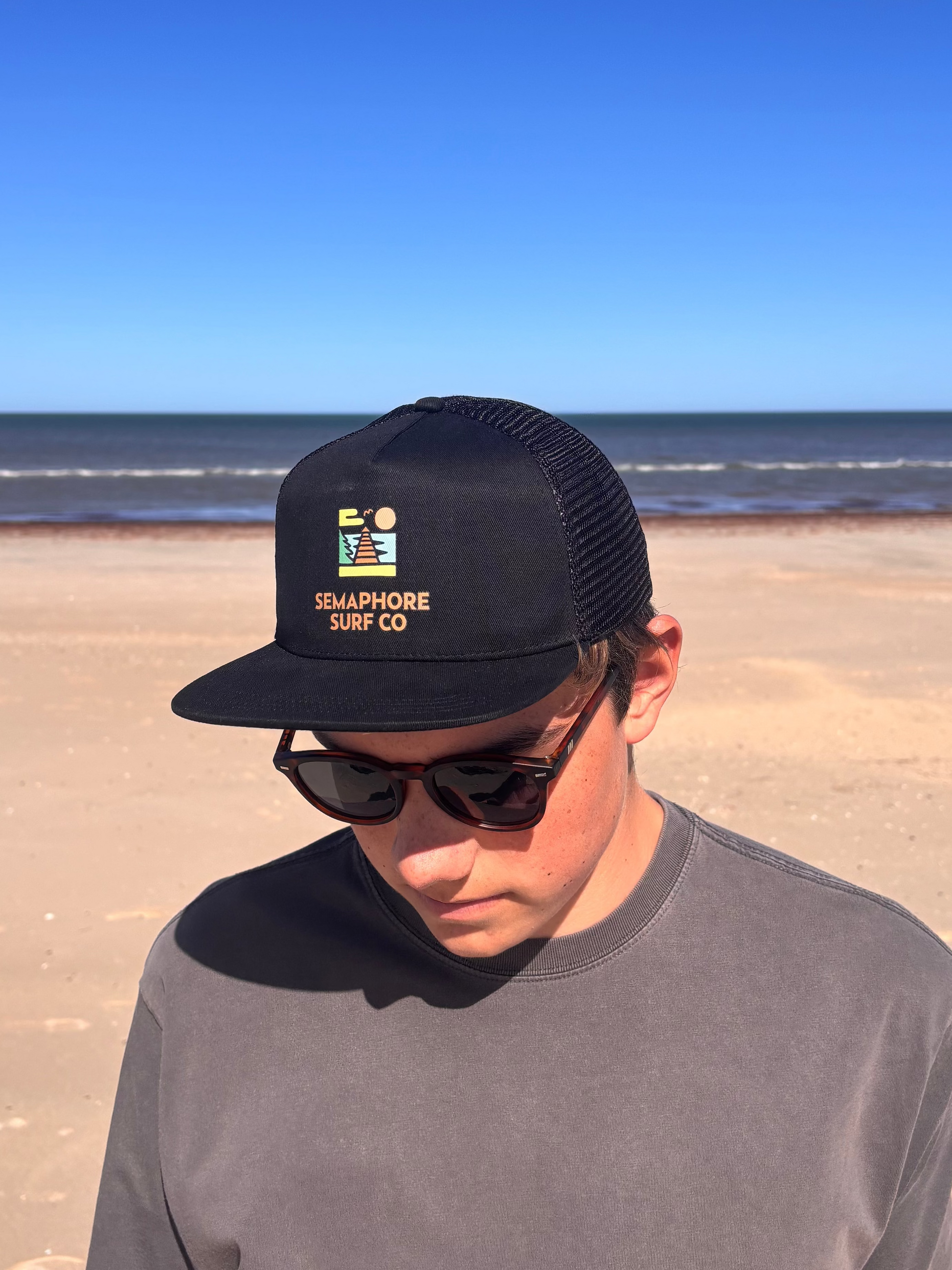 Person wearing the black Semaphore Surf Co. trucker cap and sunglasses on the beach